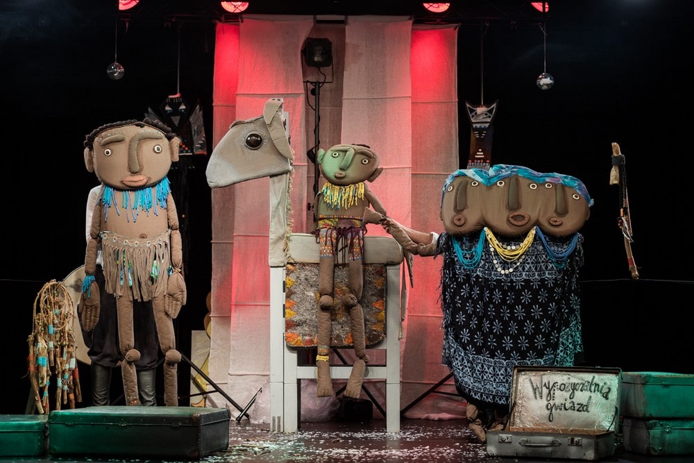 fotografia: scena z przedstawienia teatru lalkowego. stoja lalki przedstawiające w uproszczeniu ludzkie postacie. W środku jedna siedzi na figurze podobnej do konia